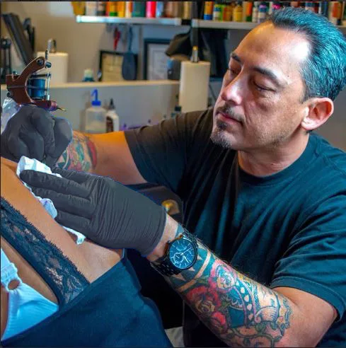 The artist in black gloves tattooing a client's shoulder, ink bottles on the shelf behind him