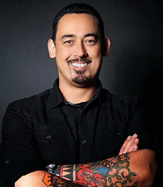 The artist smiling with arms crossed, a color tattoo sleeve showing on one forearm