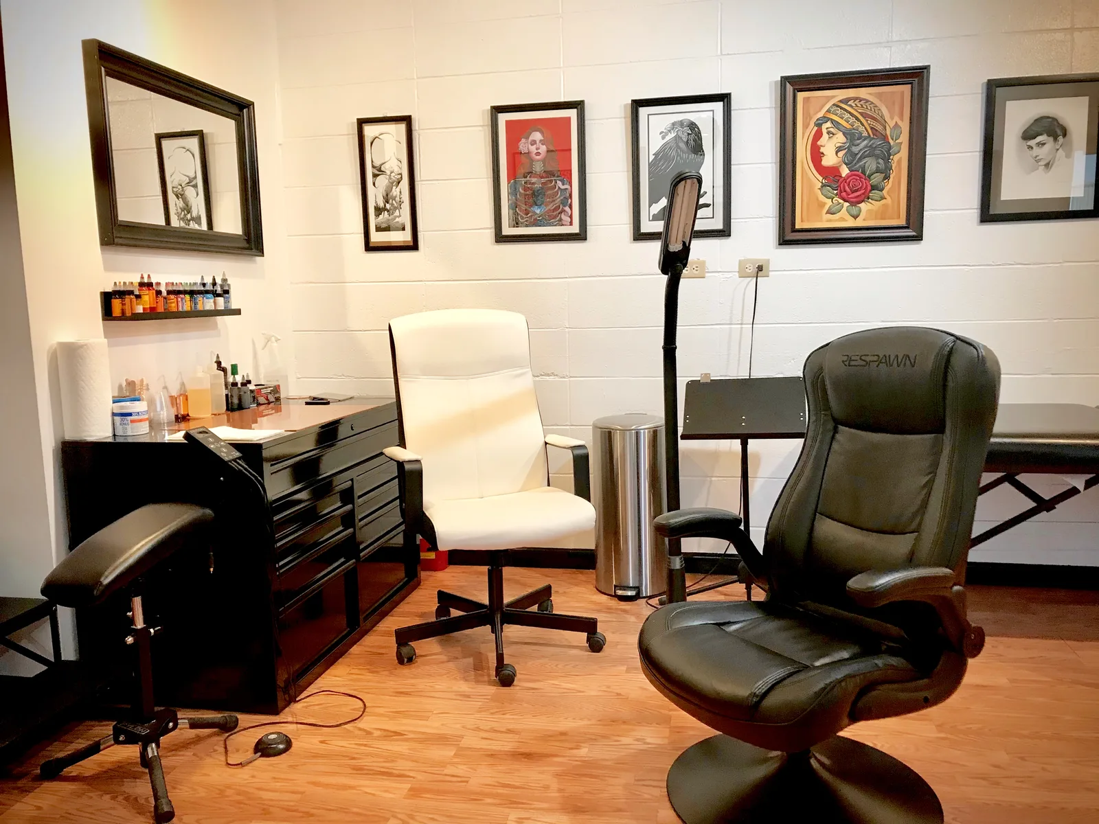 Inside the studio: a white artist chair, a black reclining client chair, a tool cabinet and framed art on white brick