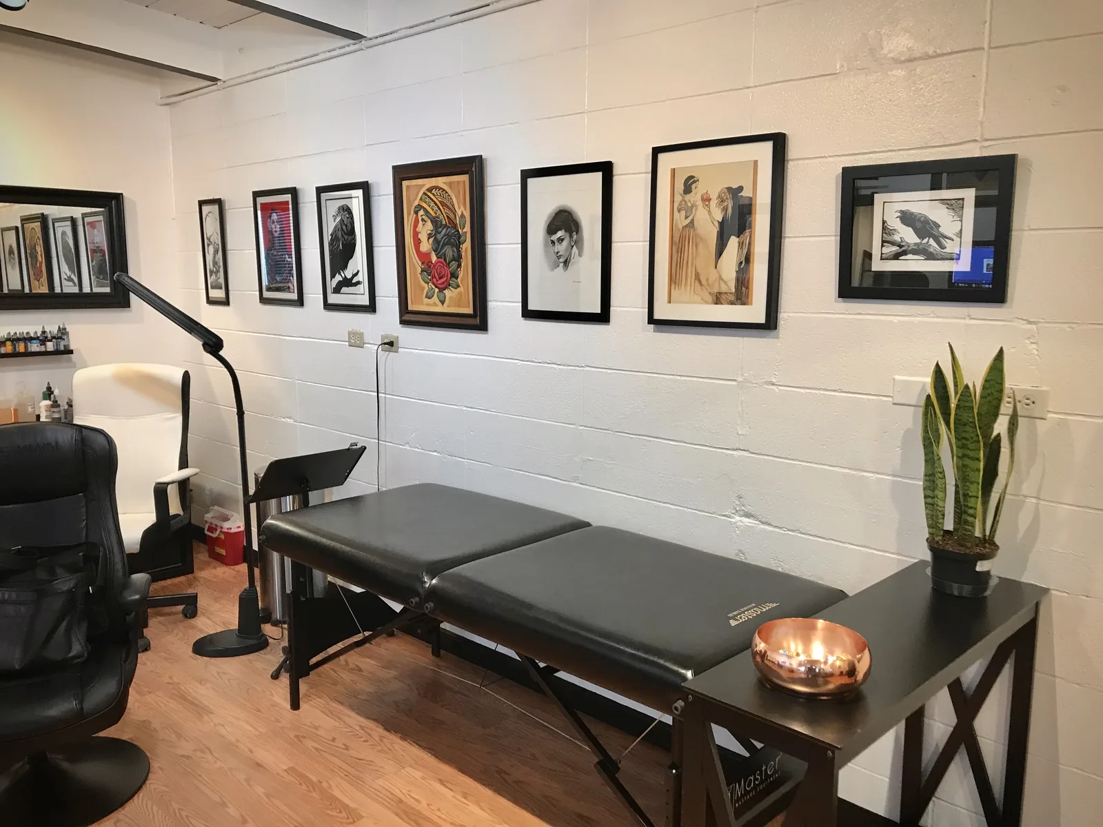 A padded black tattoo table beside a side table with a snake plant and a copper bowl of candles, under a row of framed prints